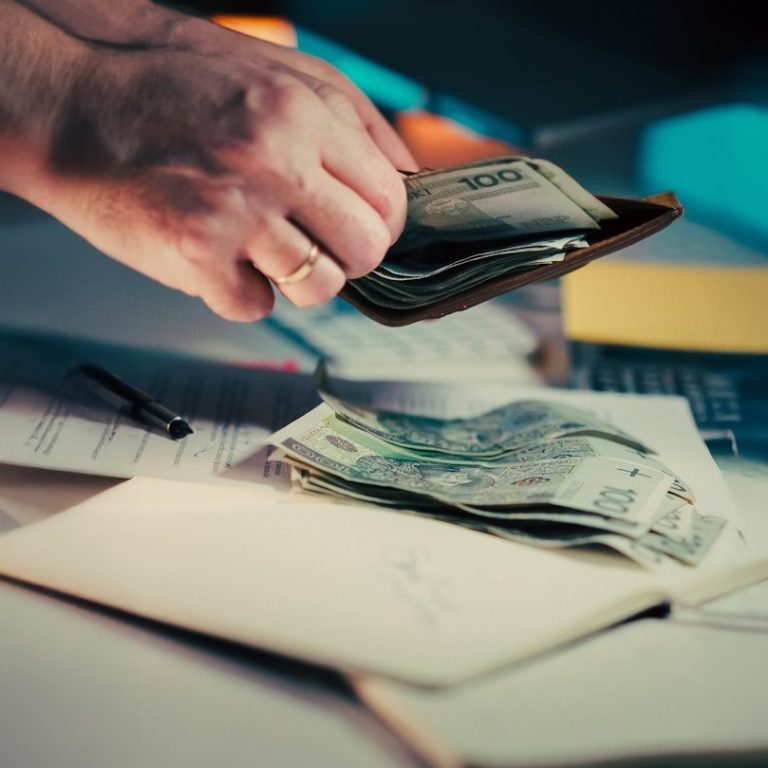 A hand holding cash above a wallet on a desk with papers and a pen.