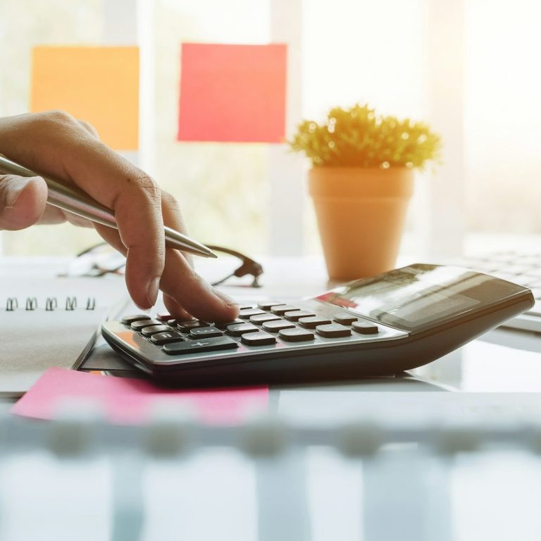 A hand using a calculator on a desk with sticky notes and a potted plant.