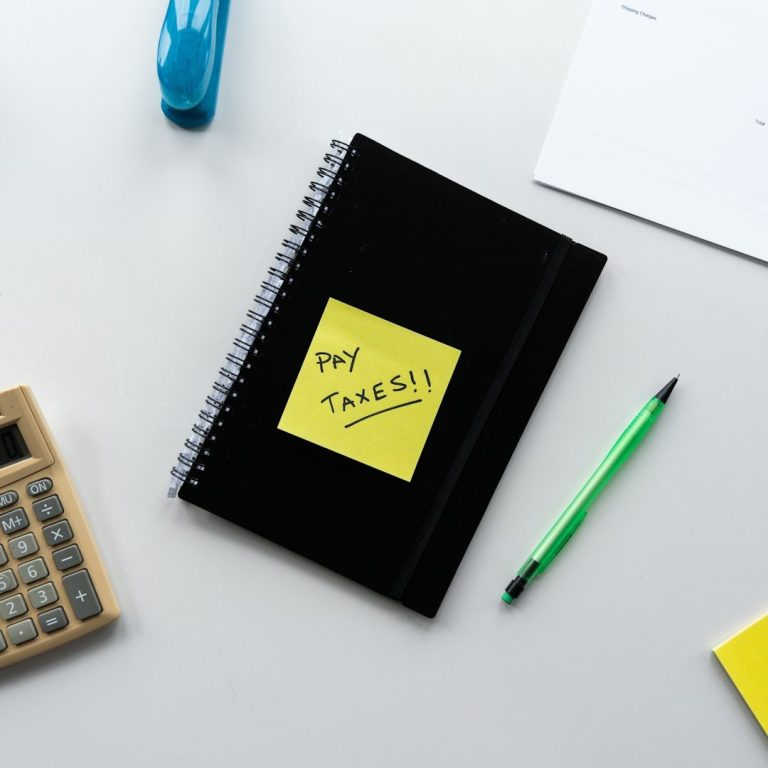 Black notebook with a yellow sticky note saying "Pay Taxes," a calculator, and a green pen.