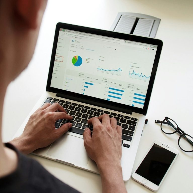 Person reviewing analytics on a laptop with graphs, a smartphone, and glasses nearby.